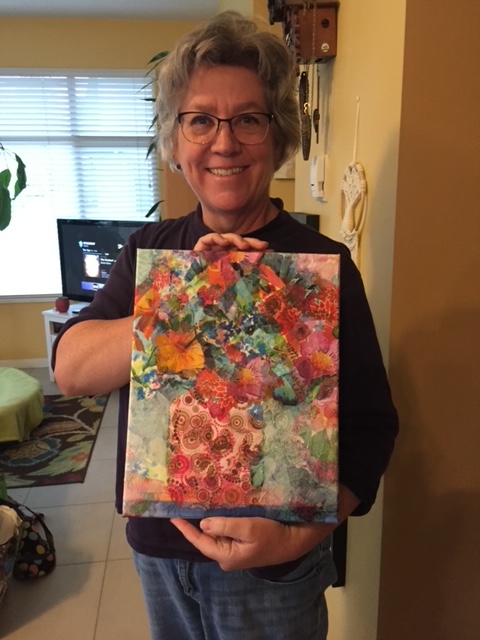 A woman holding a colorful piece of artwork featuring a floral design. The background shows a room with a window and furniture, creating a cozy atmosphere.